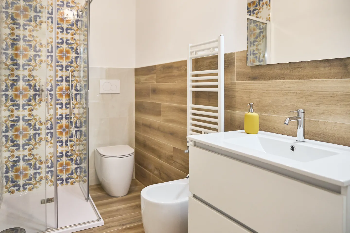 Bagno moderno dell'Appartamento Giovanna con lavabo bianco, termoarredo, bidet e doccia con rivestimento geometrico giallo e blu.